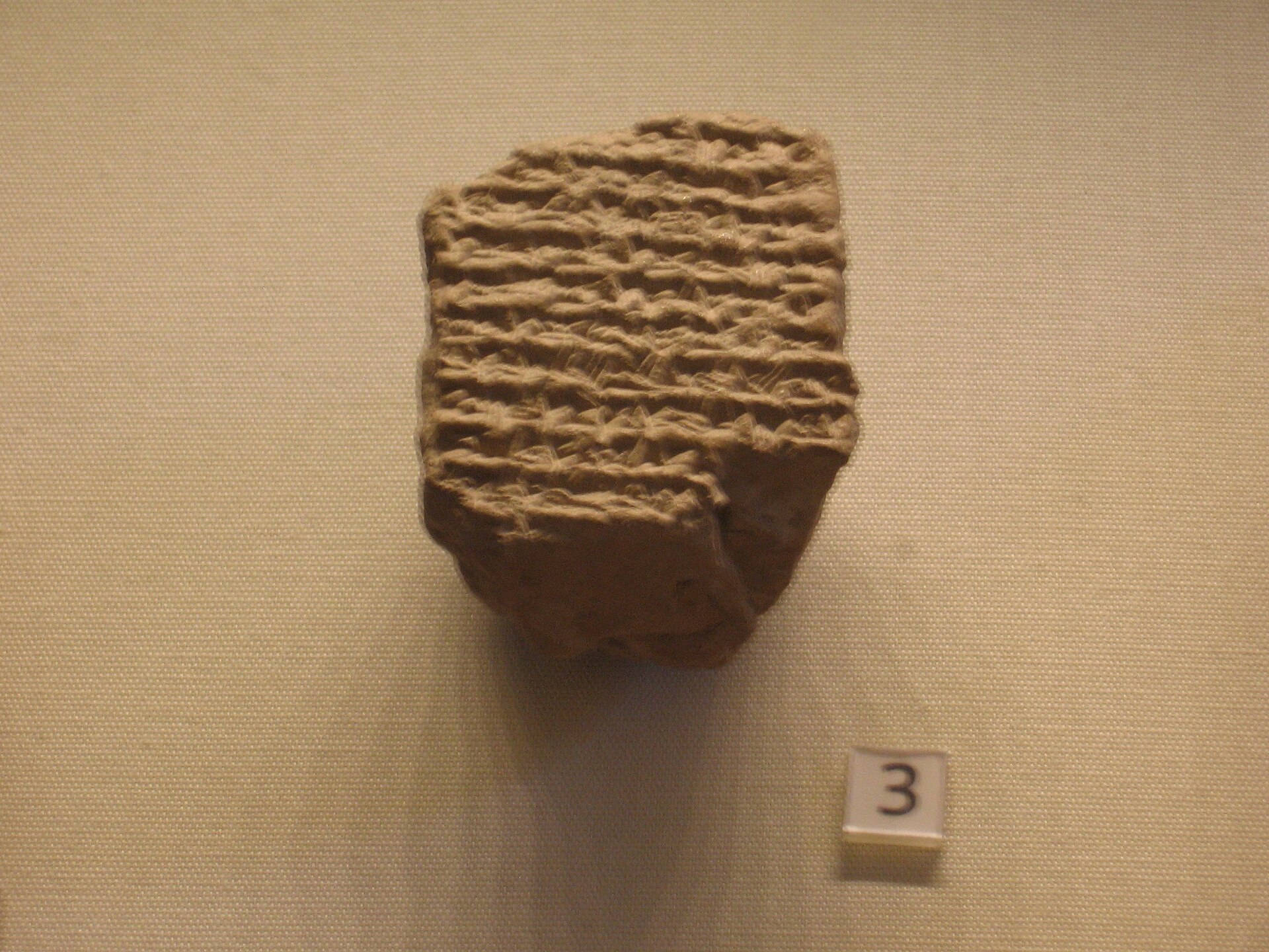 A palm-sized, broken clay tablet densely covered in cuneiform wedge-marks arranged in rows, photographed on a fabric-covered museum display board next to a small paper tag printed with the number 3.