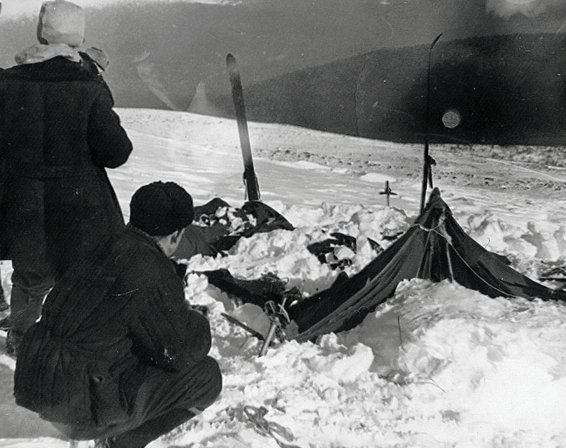 A black and white photograph of a collapsed canvas tent partly buried in snow on an open slope, with two searchers in heavy winter clothing crouched beside it and skis standing upright in the snow.
