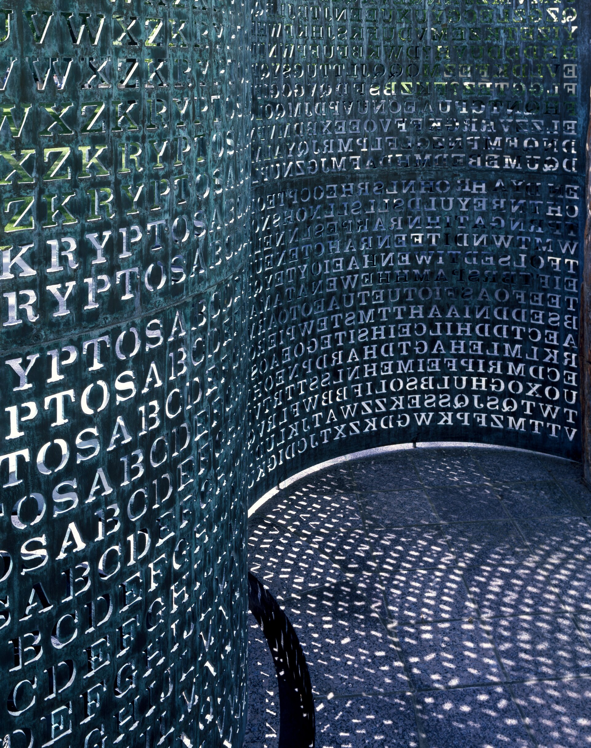 A close-up photograph of Kryptos, a curved copper sculpture with rows of letters cut through the metal. One panel shows a repeating KRYPTOS-keyed alphabet grid; the adjoining curved panel is dense with encrypted characters, their shapes projected as shadows onto the stone courtyard below.