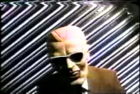 A television screen capture of a person wearing a glossy Max Headroom mask and sunglasses, in a suit jacket, standing in front of a rippling silver corrugated-metal backdrop.