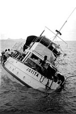 A small wooden motor vessel listing steeply to port in open water, the name JOYITA painted on her hull, with several men climbing aboard from alongside.