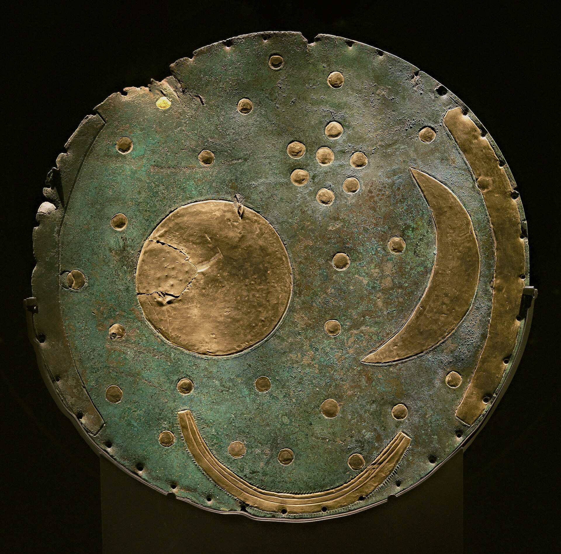 The Nebra Sky Disc, a round bronze plate corroded green with age, inlaid with a gold circle representing the sun or full moon, a gold crescent moon, a cluster of small gold dots representing stars, and a curved gold arc, photographed against a black background at the State Museum of Prehistory in Halle, Germany.