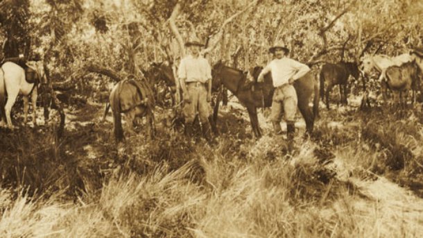 A grainy sepia photograph of two young men in expedition clothing standing with others in dense jungle brush at a campsite.