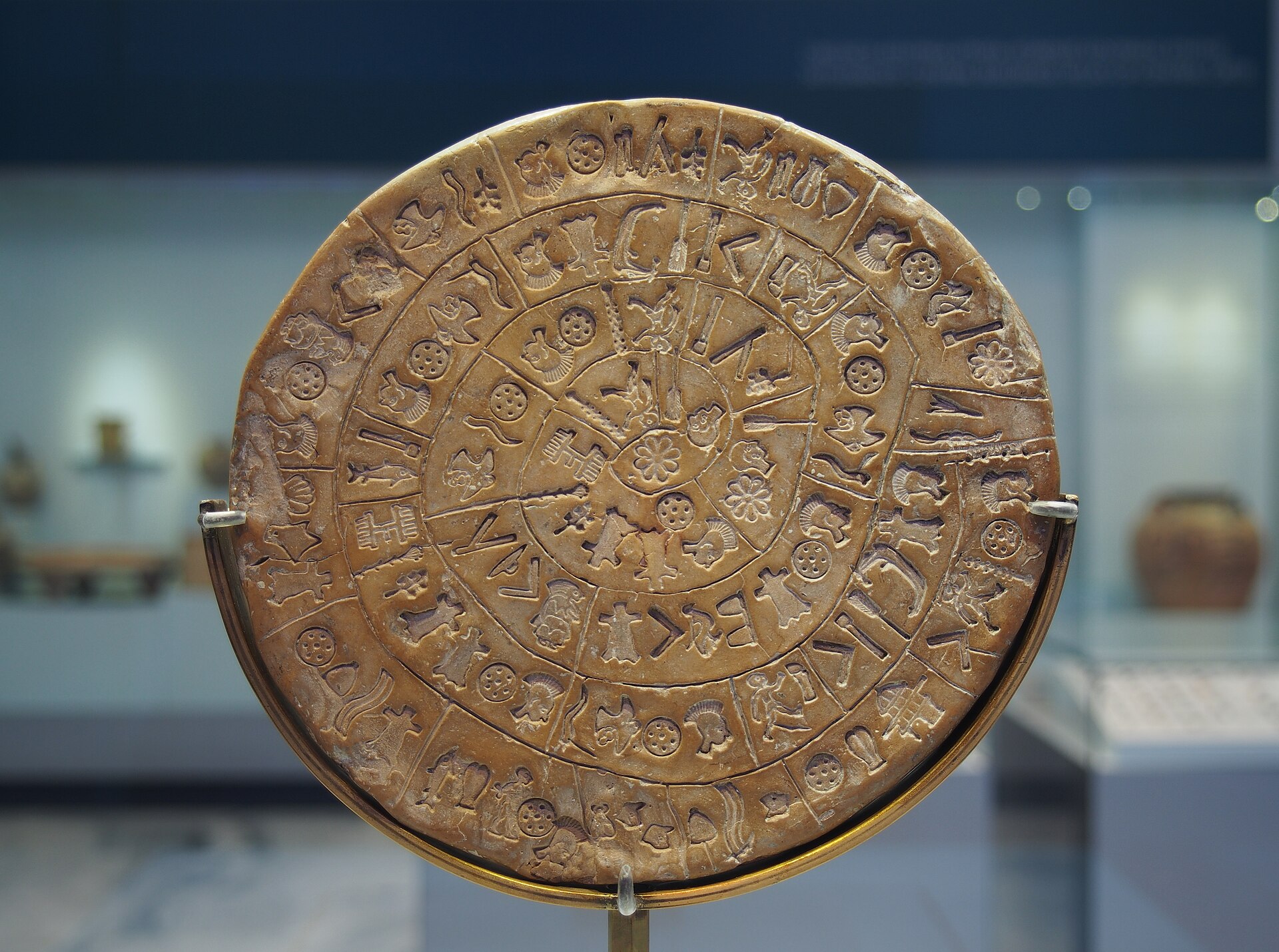 The Phaistos Disc, a round clay tablet with pictographic signs stamped in a spiral pattern across its face, mounted upright in a museum display case.