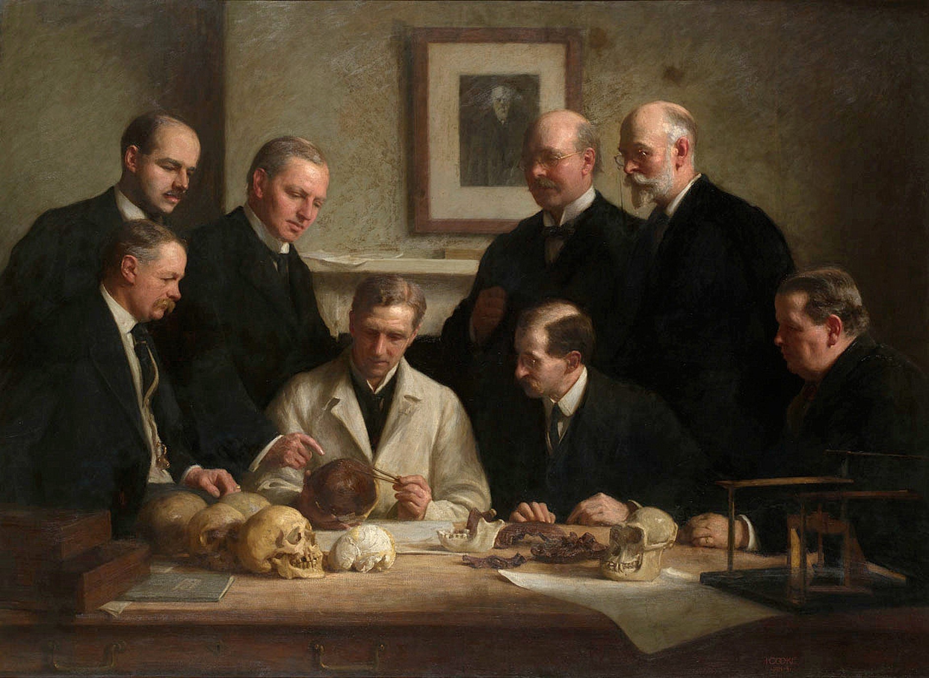 An oil painting of eight men in dark Edwardian suits gathered around a table, examining a reconstructed skull and jaw fragments laid out among other skulls and bones, with a framed portrait hanging on the wall behind them.