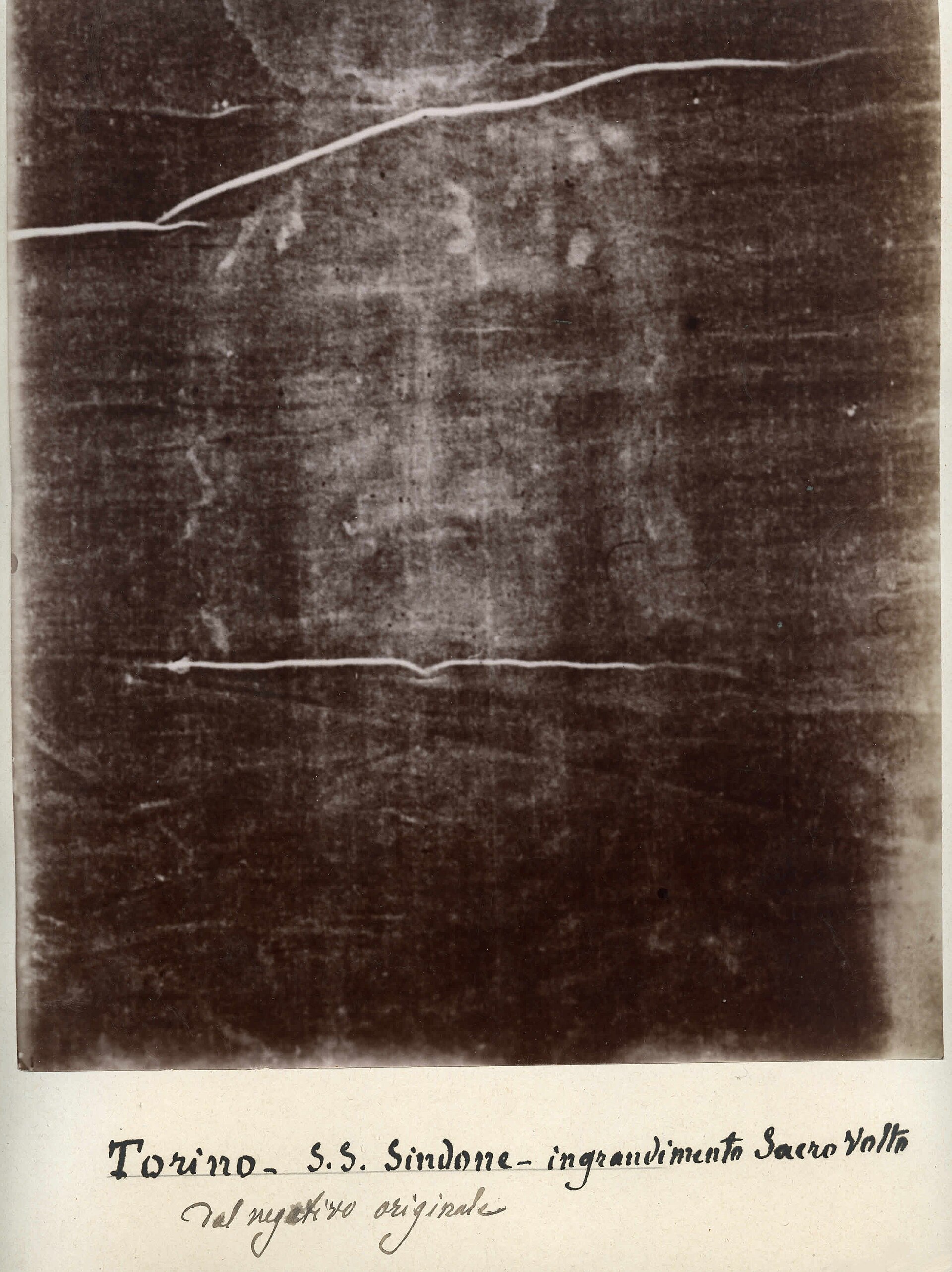 A sepia-toned photographic print mounted on aged card, showing a faint, ghostly image of a bearded man's face and shoulders rendered as light tones against a dark ground, with handwritten Italian text below reading 'Torino - S.S. Sindone - ingrandimento Sacro Volto / dal negativo originale.'